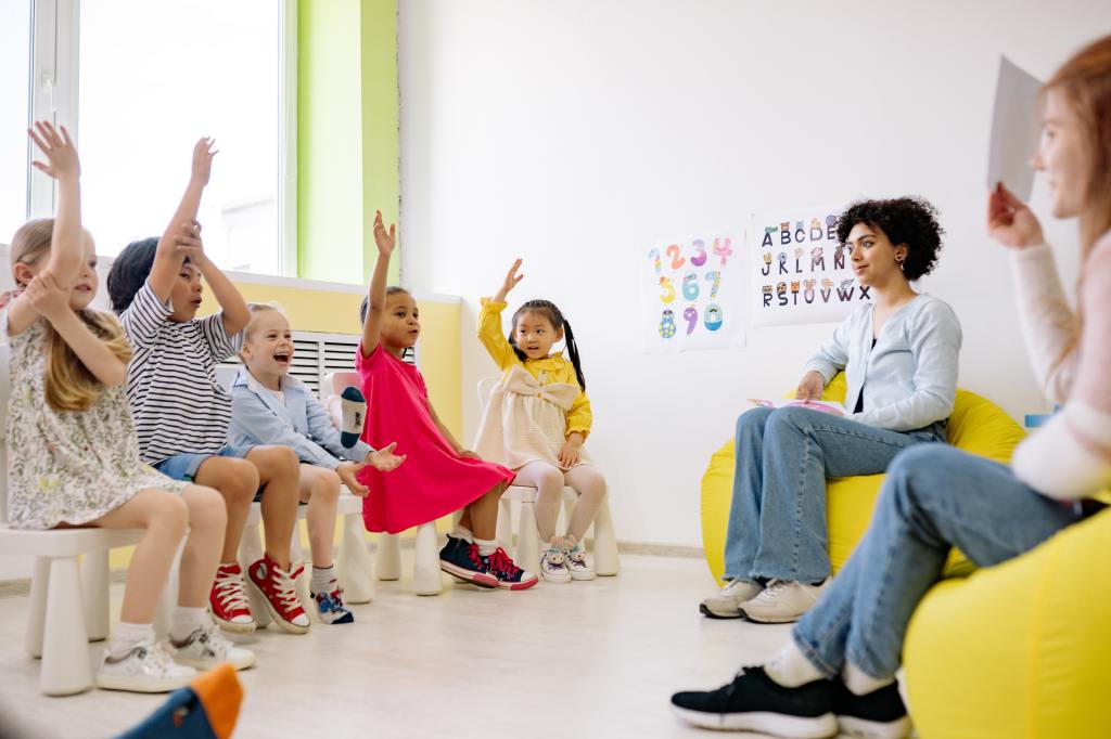 A group of kids with their hands up sit in front of two daycare workers in a daycare setting.